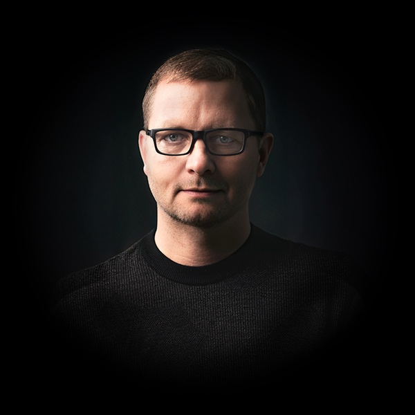 Mann mit Brille in dunklem Pullover, vor dunklem Hintergrund.